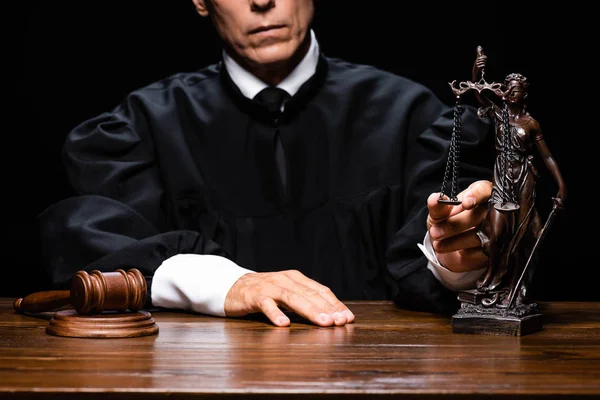 Cropped view of judge in judicial robe sitting at table with gavel and themis figure isolated on black — Stock Photo
