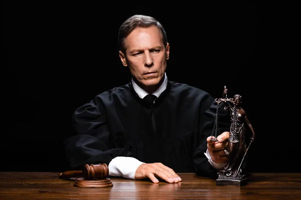 Judge in judicial robe sitting at table with gavel and themis figure isolated on black — Stock Photo