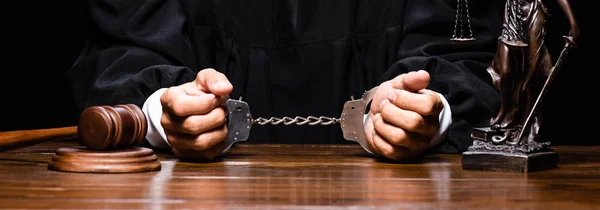 Panoramic shot of judge in judicial robe sitting at table with handcuffs isolated on black — Stock Photo