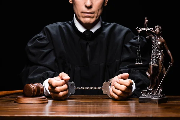 Обрізаний вид судді в судовому халаті, сидячи за столом з наручниками ізольовані на чорному — стокове фото