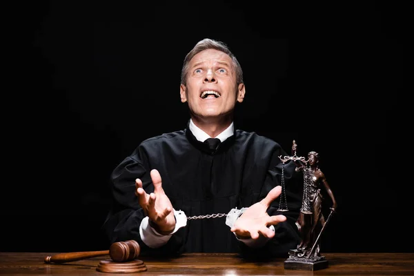Angry judge in judicial robe sitting at table with handcuffs and scream isolated on black — Stock Photo