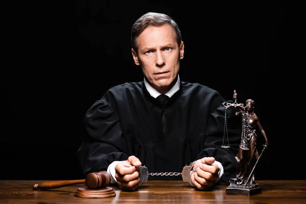 Judge in judicial robe sitting at table with handcuffs isolated on black — Stock Photo