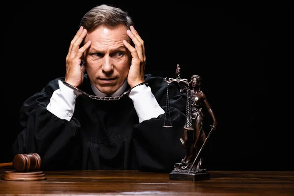 Judge in judicial robe sitting at table with handcuffs isolated on black — Stock Photo