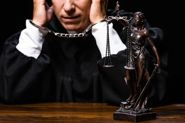 Обрезанный взгляд судьи в судейском халате, сидящего за столом с наручниками, изолированными на черном — стоковое фото