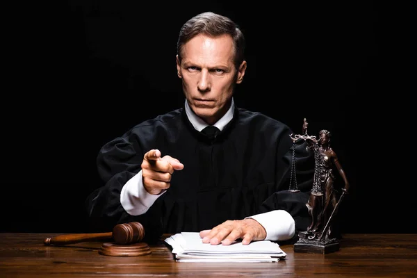 Juez en bata judicial sentado en la mesa y señalando con el dedo aislado en negro - foto de stock