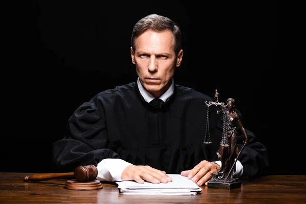 Judge in judicial robe sitting at table and looking at camera isolated on black — Stock Photo