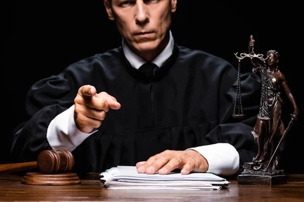 Vista recortada del juez en bata judicial sentado a la mesa y señalando con el dedo aislado en negro - foto de stock