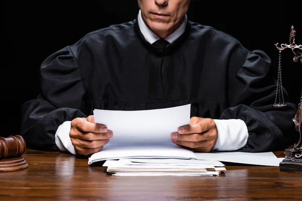 Cropped view of judge in judicial robe sitting at table and reading paper isolated on black — Stock Photo