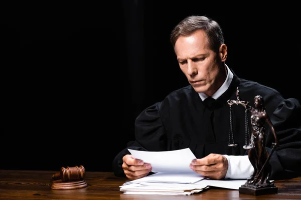 Judge in judicial robe sitting at table and reading paper isolated on black — Stock Photo