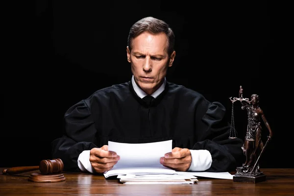 Judge in judicial robe sitting at table and reading paper isolated on black — Stock Photo