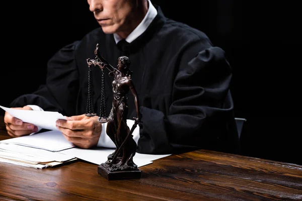 Vista recortada del juez en bata judicial sentado en la mesa y papel de lectura aislado en negro - foto de stock