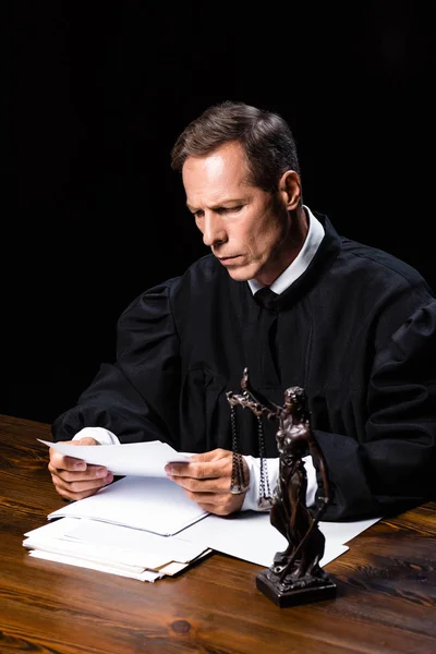 Judge in judicial robe sitting at table and reading paper isolated on black — Stock Photo