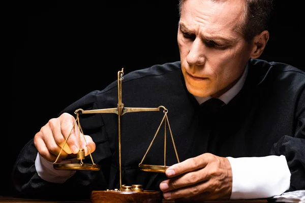 Judge in judicial robe sitting at table and holding scales of justice isolated on black — Stock Photo