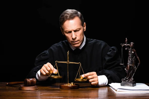 Judge in judicial robe sitting at table and holding scales of justice isolated on black — Stock Photo