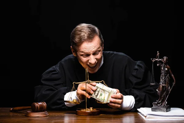 Juez sonriente en bata judicial sentado a la mesa y sosteniendo billetes de dólar aislados en negro - foto de stock