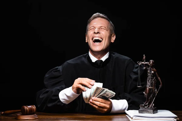 Juez sonriente en bata judicial sentado a la mesa y sosteniendo billetes de dólar aislados en negro - foto de stock