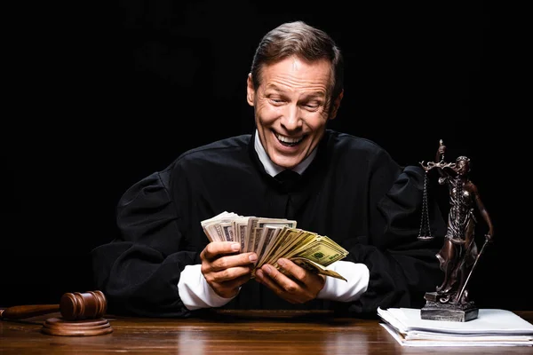 Juez sonriente en bata judicial sentado a la mesa y sosteniendo billetes de dólar aislados en negro - foto de stock