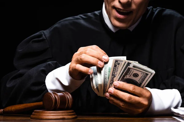 Ausgeschnittene Ansicht eines schockierten Richters in Richterrobe, der am Tisch sitzt und Dollarnoten isoliert auf schwarzem Grund zählt — Stockfoto