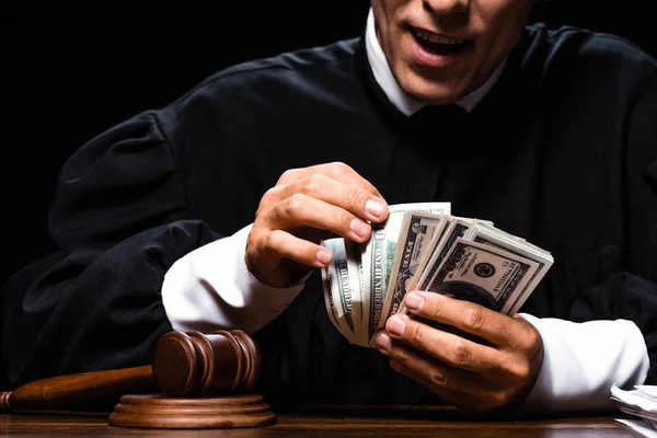 Cropped view of shocked judge in judicial robe sitting at table and counting dollar banknotes isolated on black — Stock Photo
