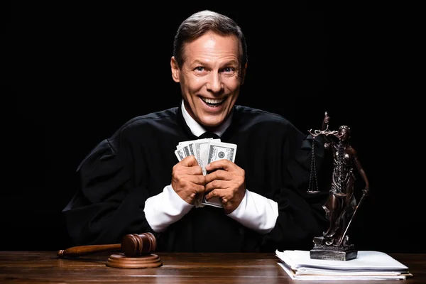 Juez sonriente en bata judicial sentado a la mesa y sosteniendo billetes de dólar aislados en negro - foto de stock