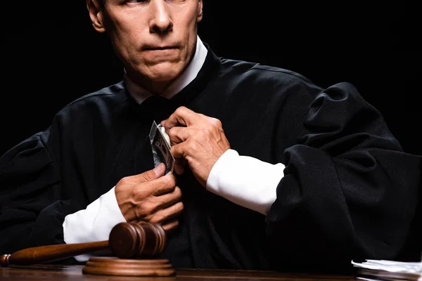 Обрізаний вид на наляканого судді в судовому халаті, що сидить за столом і тримає доларові банкноти ізольовані на чорному — стокове фото