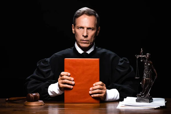 Giudice in veste giudiziaria seduto a tavola e tenendo libro arancione isolato su nero — Foto stock