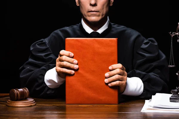 Vista recortada del juez en bata judicial sentado a la mesa y sosteniendo libro naranja aislado en negro - foto de stock