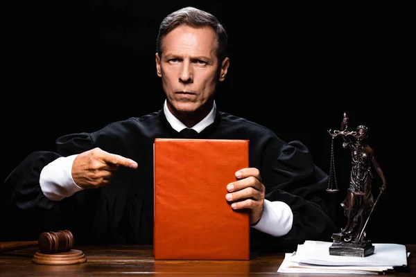 Суддя в судовому халаті сидить за столом і вказує пальцем на помаранчеву книгу ізольовано на чорному — стокове фото
