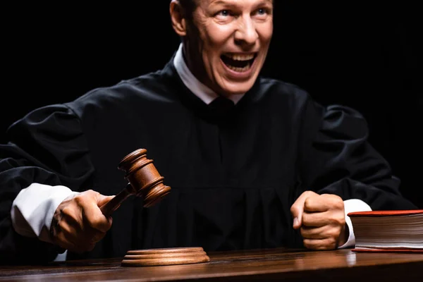 Angry judge in judicial robe sitting at table and hitting with gavel isolated on black — Stock Photo