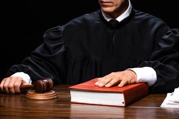 Corte vista do juiz em roupão judicial sentado à mesa e segurando martelo e livro isolado em preto — Fotografia de Stock