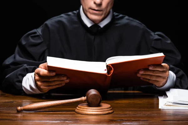 Vista recortada del juez en bata judicial sentado a la mesa y leyendo libro naranja aislado en negro - foto de stock