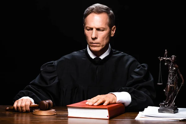 Judge in judicial robe holding gavel and putting hand on book isolated on black — Stock Photo