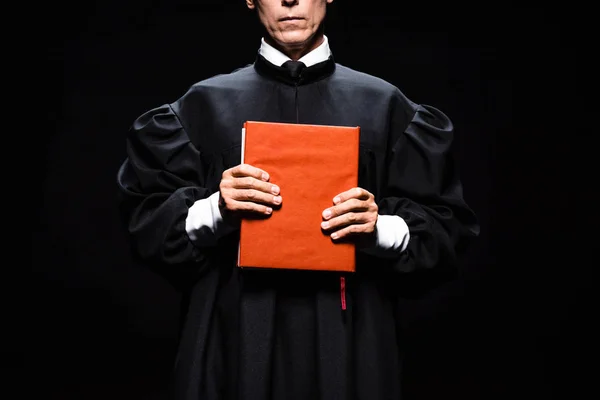 Cropped view of judge in judicial robe holding orange book isolated on black — Stock Photo