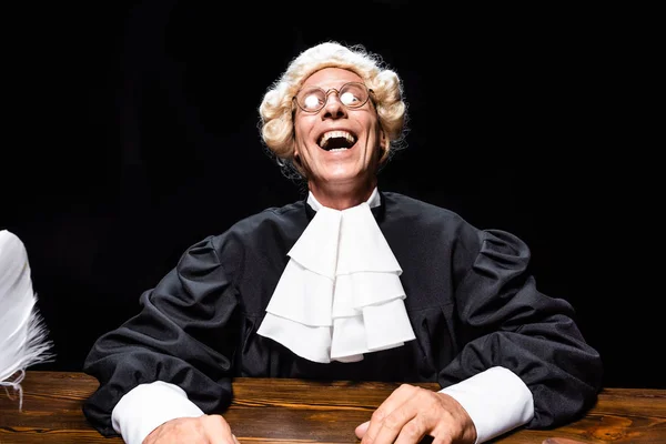 Sonriente juez en bata judicial y peluca sentado a la mesa aislado en negro - foto de stock