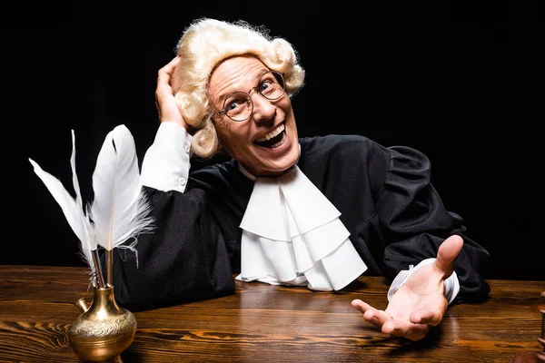 Улыбающийся судья в судейском халате и парике, сидящий за столом изолированный на черном — стоковое фото