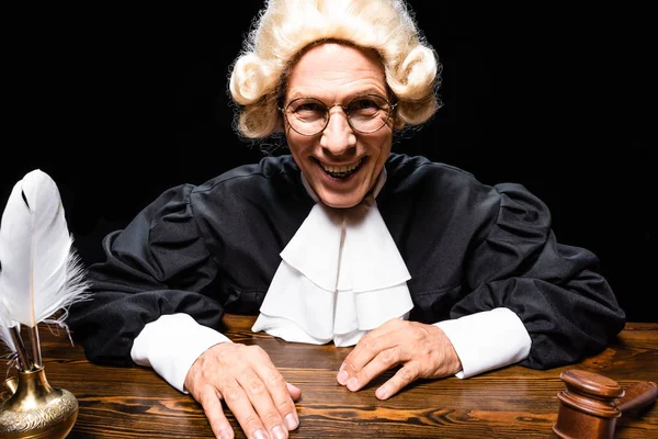 Sorrindo juiz em roupão judicial e peruca sentado à mesa isolado em preto — Fotografia de Stock