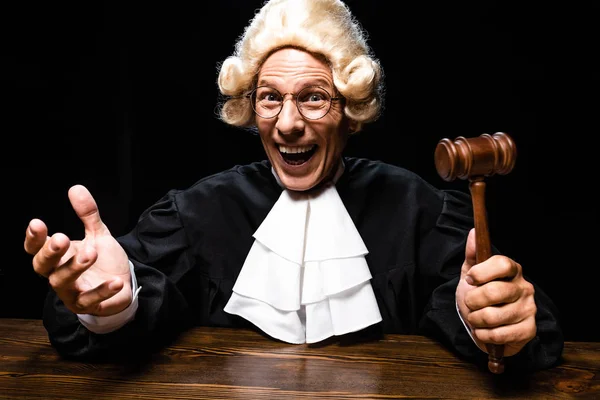 Smiling judge in judicial robe and wig sitting at table and holding gavel isolated on black — Stock Photo