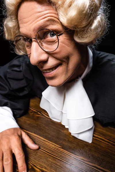 Sonriente juez en bata judicial y peluca sentado a la mesa aislado en negro - foto de stock