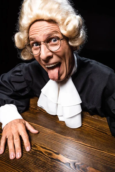 Judge in judicial robe and wig sitting at table and sticking out tongue isolated on black — Stock Photo