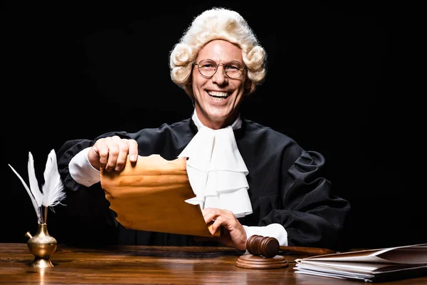 Juge souriant en robe judiciaire et perruque assis à la table et tenant le document isolé sur noir — Photo de stock