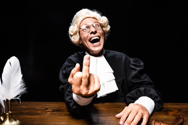 Sorrindo juiz em roupão judicial e peruca sentado à mesa e mostrando dedo médio isolado em preto — Fotografia de Stock