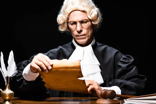 Judge in judicial robe and wig sitting at table and reading document isolated on black — Stock Photo