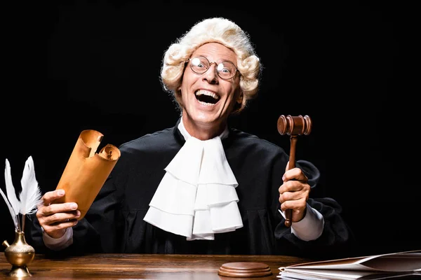 Juge souriant en robe judiciaire et perruque assis à la table et tenant le document et le marteau isolé sur noir — Photo de stock