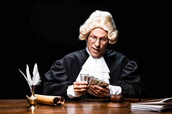 Judge in judicial robe and wig sitting at table and counting money isolated on black — Stock Photo