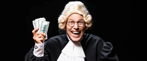 Plano panorámico de sonriente juez en bata judicial y peluca sosteniendo el dinero aislado en negro - foto de stock