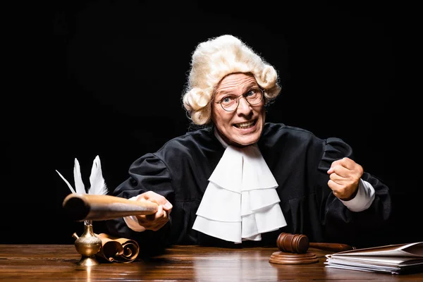 Angry judge in judicial robe and wig sitting at table and holding bat isolated on black — Stock Photo