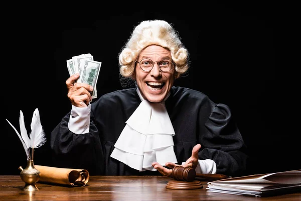 Sonriente juez en bata judicial y peluca sentado a la mesa y sosteniendo el dinero aislado en negro - foto de stock