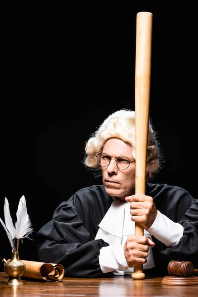 Judge in judicial robe and wig sitting at table and holding bat isolated on black — Stock Photo