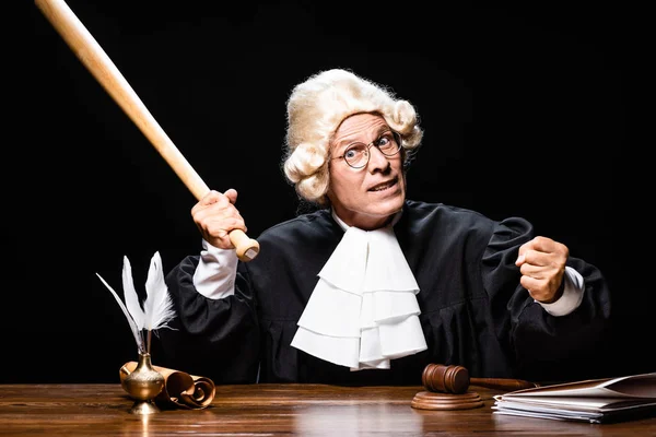 Angry judge in judicial robe and wig sitting at table and holding bat isolated on black — Stock Photo