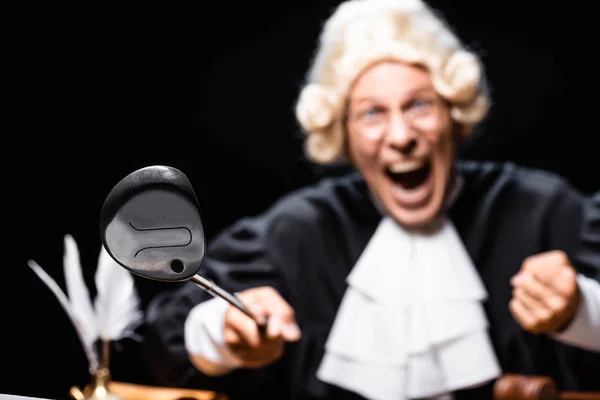 Selective focus of angry judge in judicial robe and wig holding golf club isolated on black — Stock Photo